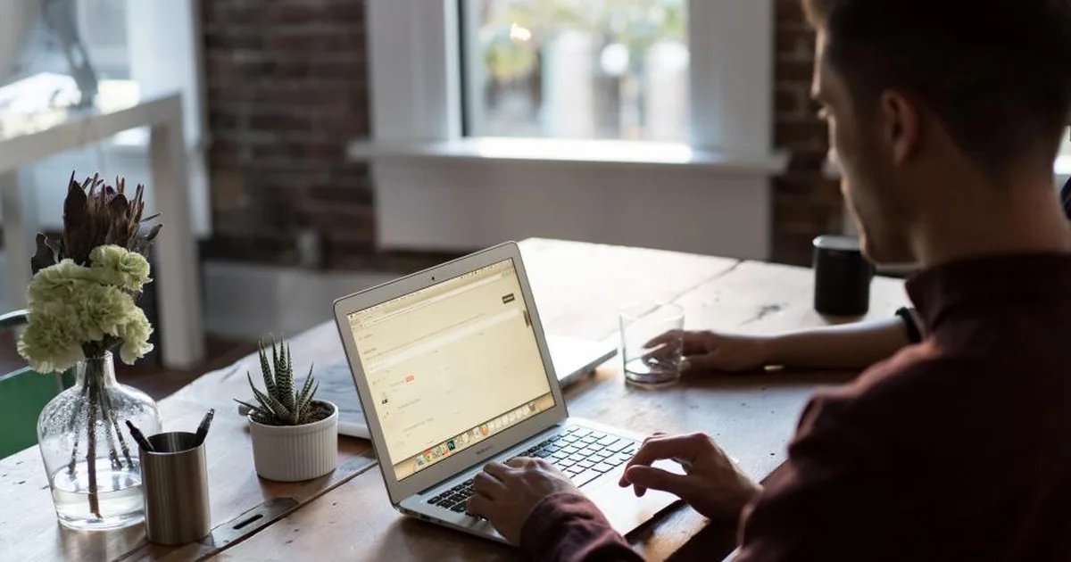 Hombre trabajando en una laptop sobre una mesa de madera junto a una ventana