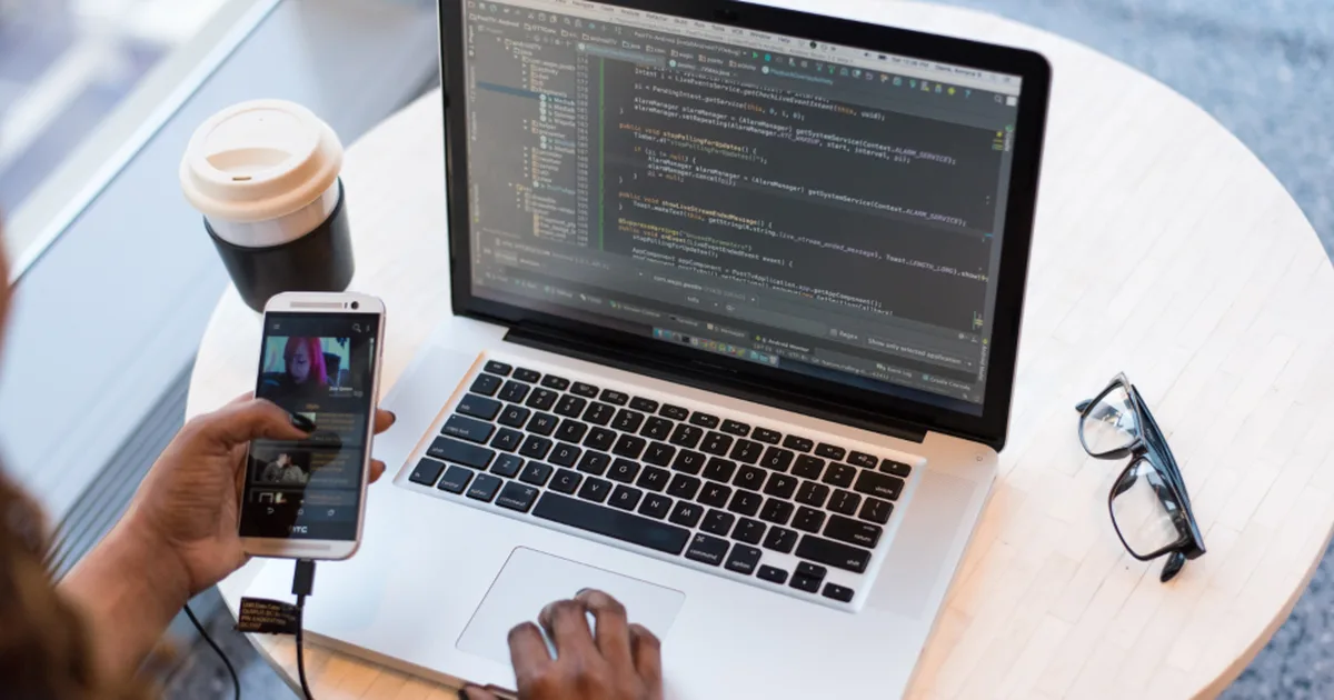 Desarrolladora sosteniendo un celular frente a una laptop con código en pantalla